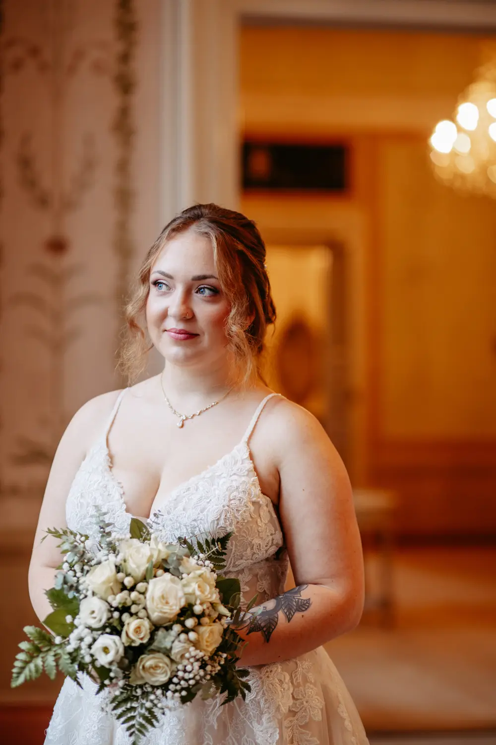 Eine Frau in einem weißen Spitzenhochzeitskleid hält einen Strauß weißer und cremefarbener Blumen. Sie steht in einem Innenraum mit sanfter Beleuchtung und eleganter Dekoration im Hintergrund. Sie hat hellbraunes Haar, das mit lockeren Locken frisiert ist.
