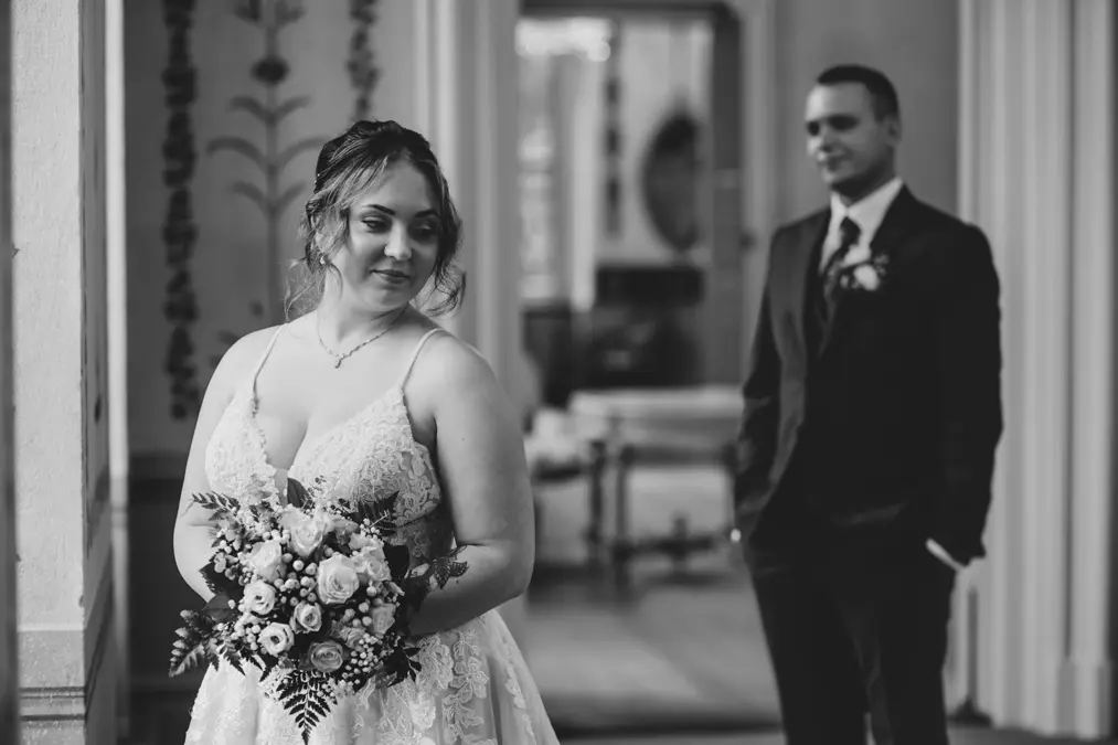 Eine Braut in einem weißen Spitzenkleid, die einen Blumenstrauß hält, steht im Vordergrund, blickt nach unten und lächelt, während der Bräutigam in einem dunklen Anzug unscharf im Hintergrund steht. Das Bild ist in schwarz-weiß gehalten.