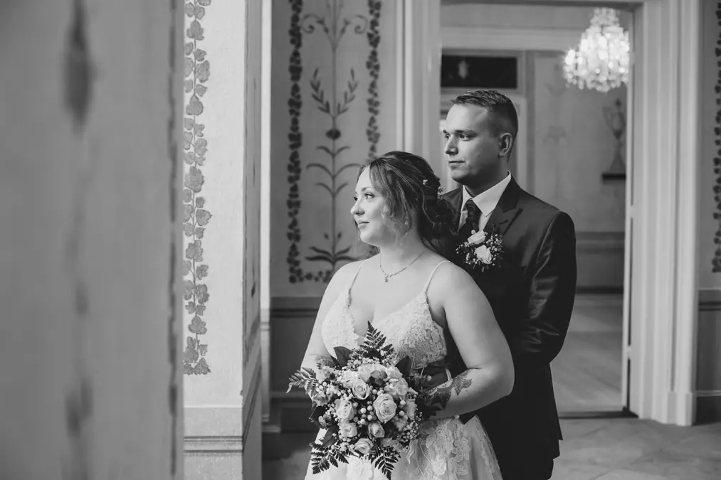 Eine Braut und ein Bräutigam stehen zusammen in einem Haus und blicken aus einem Fenster. Die Braut hält einen Blumenstrauß und trägt ein Spitzenkleid, während der Bräutigam in Anzug und Krawatte hinter ihr steht. Das Bild ist in schwarz-weiß gehalten.