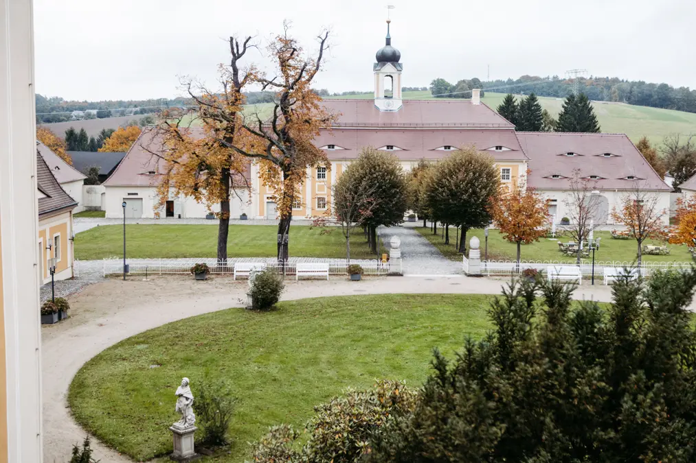 Ein großes Gebäude mit rotem Dach und zentralem Uhrenturm befindet sich hinter einem runden grasbewachsenen Innenhof mit Bäumen, Bänken und einer Statue; im Hintergrund sind Herbstlaub und sanfte Hügel zu sehen.