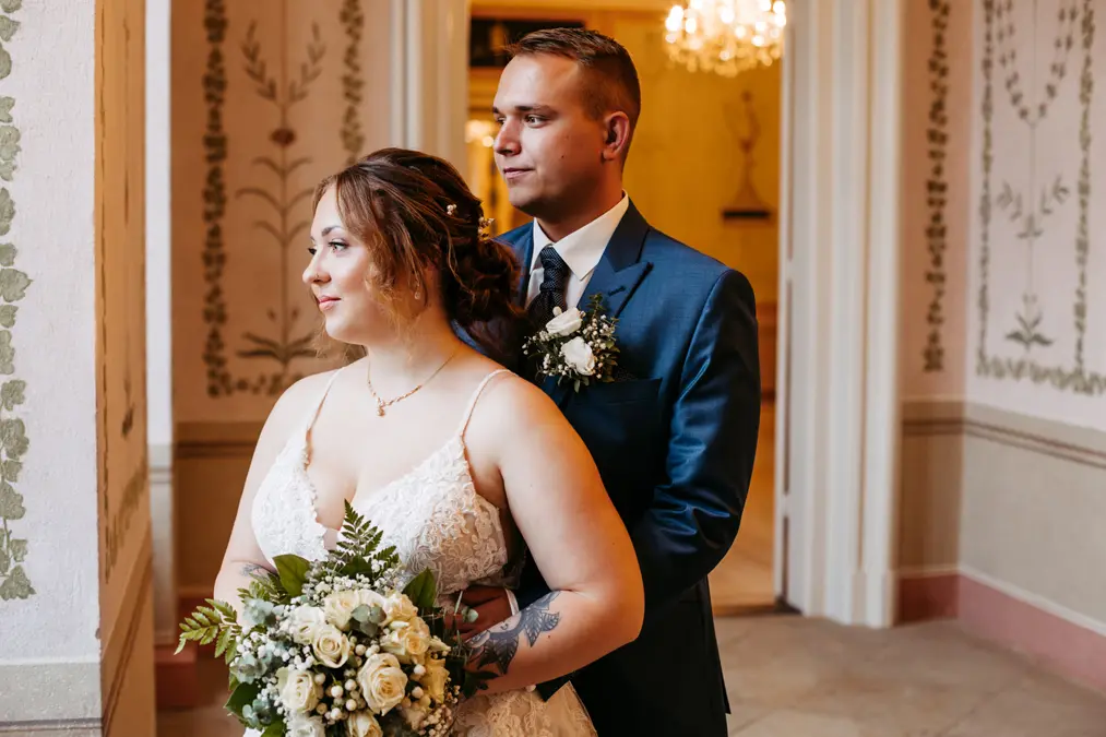 Eine Braut in einem weißen Kleid mit einem Blumenstrauß in der Hand steht vor einem Bräutigam in einem blauen Anzug. Sie befinden sich in einem Innenraum, umgeben von elegantem Blumenschmuck an der Wand, und beide sehen zufrieden und nachdenklich aus.