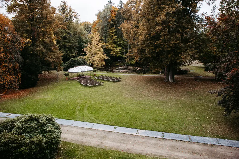 Eine Hochzeit im Freien auf einem grasbewachsenen Rasen mit Stuhlreihen vor einem weißen Baldachin, umgeben von hohen Bäumen mit Herbstlaub in einer friedlichen Parklandschaft.