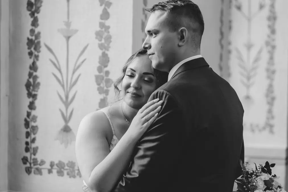 Die Braut legt ihren Kopf mit geschlossenen Augen auf die Brust des Bräutigams, während dieser nach vorne blickt. Sie stehen dicht beieinander in einem dekorierten Raum mit Blumenmustern an den Wänden. Das Foto ist in schwarz-weiß gehalten.