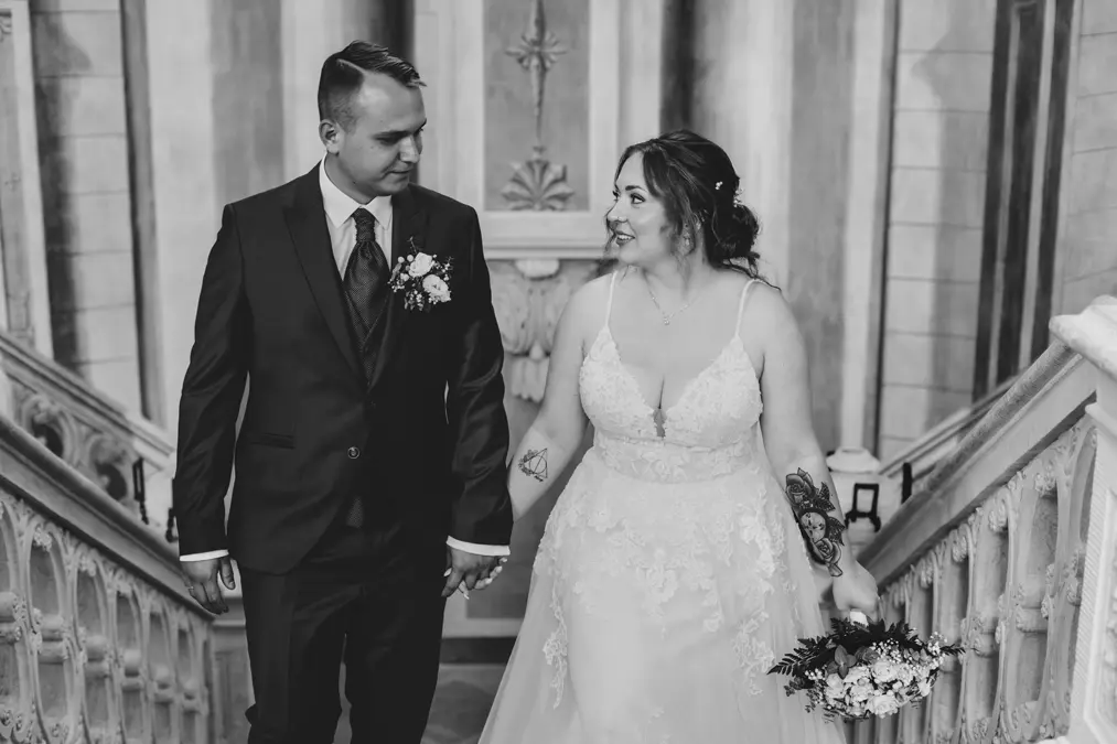 Eine Braut und ein Bräutigam halten sich an den Händen und lächeln sich an, während sie auf einer Treppe stehen. Die Braut trägt ein Spitzenkleid und hat einen Blumenstrauß in der Hand; der Bräutigam trägt einen dunklen Anzug und eine Ansteckblume. Das Foto ist in schwarz-weiß gehalten.