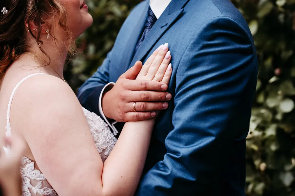 Eine Braut und ein Bräutigam stehen dicht beieinander; die Hand der Braut liegt auf der Brust des Bräutigams und zeigt ihre Eheringe. Der Bräutigam trägt einen blauen Anzug, und das Grün im Hintergrund ist unscharf.