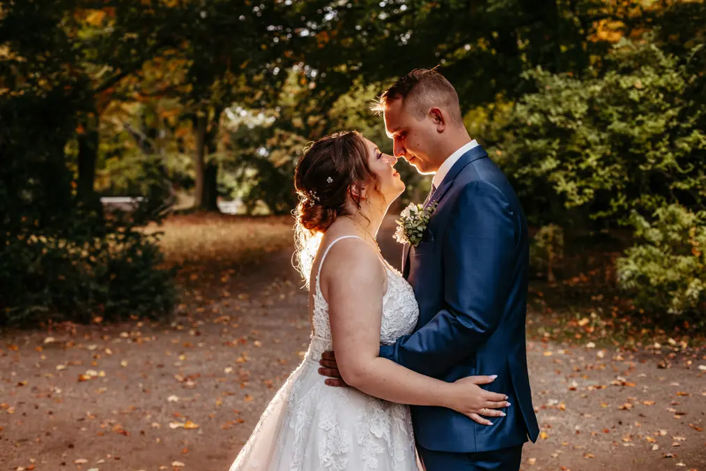 Eine Braut in einem weißen Kleid und ein Bräutigam in einem blauen Anzug stehen im Freien dicht beieinander, blicken sich in die Augen und lächeln, umgeben von Bäumen mit Herbstlaub.
