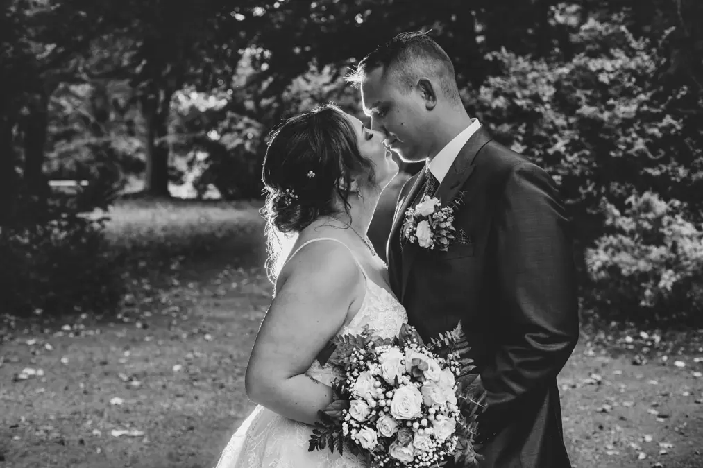 Eine Braut und ein Bräutigam stehen im Freien dicht beieinander, lächeln und berühren sich an der Stirn. Die Braut hält einen Blumenstrauß in der Hand. Das Schwarz-Weiß-Foto fängt einen zarten, romantischen Moment inmitten von Bäumen und Laub ein.