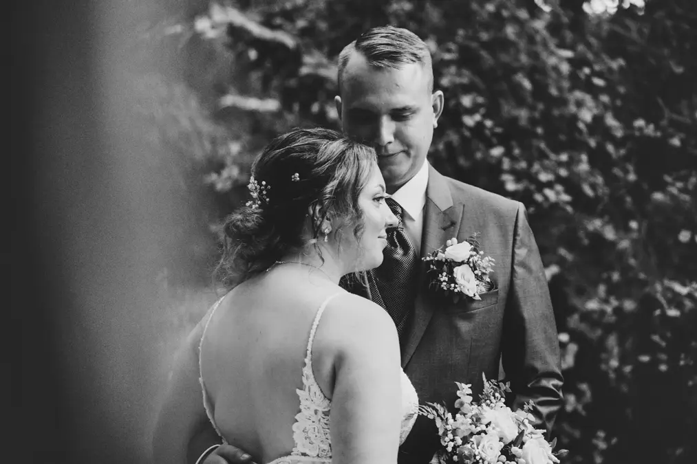 Eine Braut und ein Bräutigam umarmen sich im Freien und schauen sich liebevoll an. Der Bräutigam trägt einen Anzug mit einer Ansteckblume, die Braut ein Spitzenkleid und hält einen Blumenstrauß in der Hand. Das Foto ist schwarz-weiß und hat einen natürlichen, blattreichen Hintergrund.