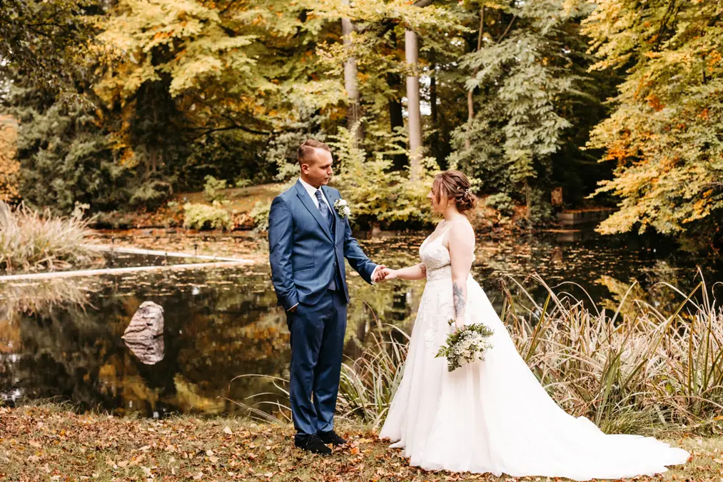 Eine Braut in einem weißen Kleid und mit einem Blumenstrauß in der Hand steht händchenhaltend mit einem Bräutigam in einem blauen Anzug an einem Teich, umgeben von hohen Bäumen mit grünen und gelben Blättern.