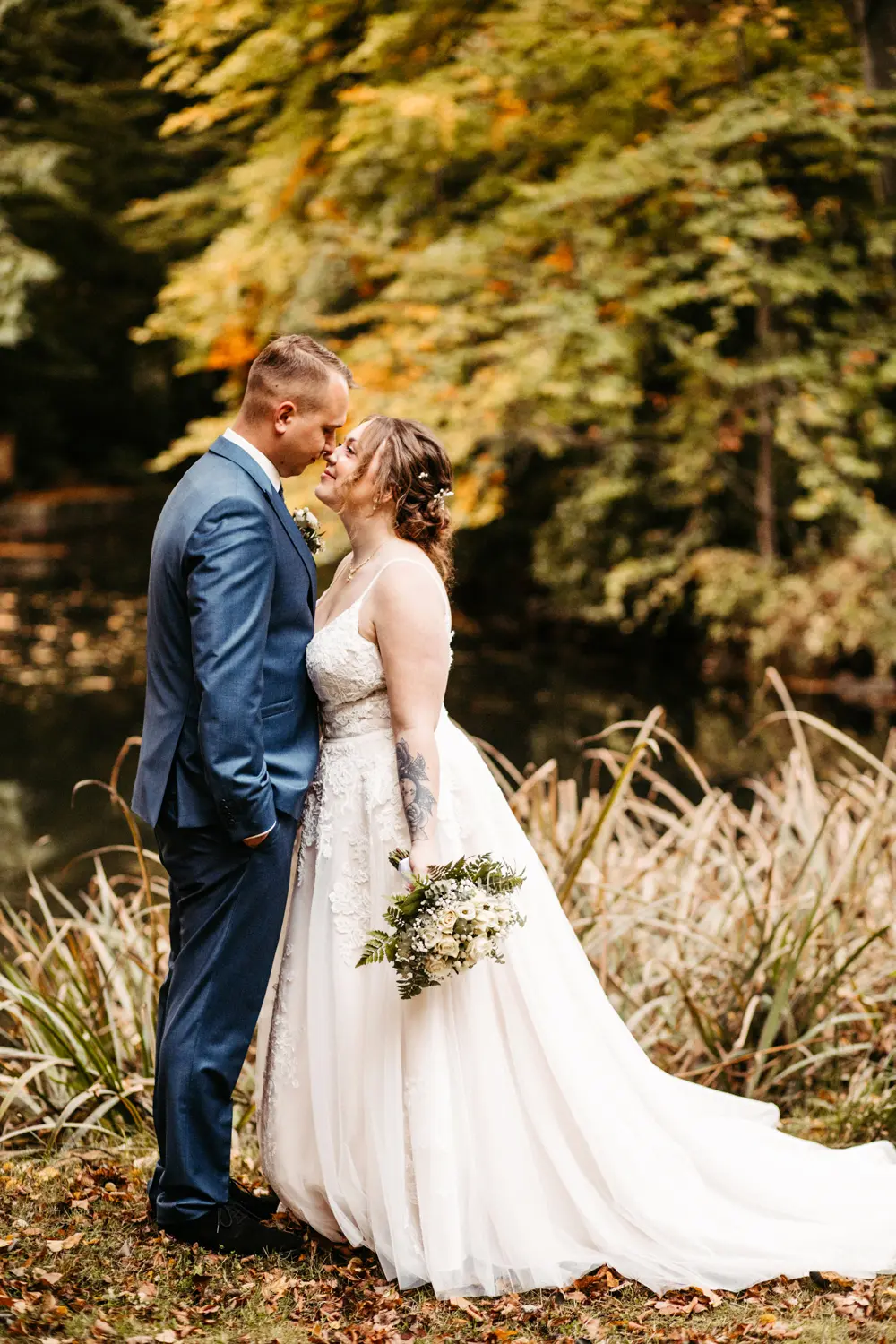 Eine Braut und ein Bräutigam stehen dicht beieinander im Freien, einander zugewandt und lächelnd, umgeben von Grün und herbstlich gefärbten Bäumen. Die Braut hält einen Blumenstrauß in der Hand und trägt ein weißes Kleid, der Bräutigam trägt einen blauen Anzug.
