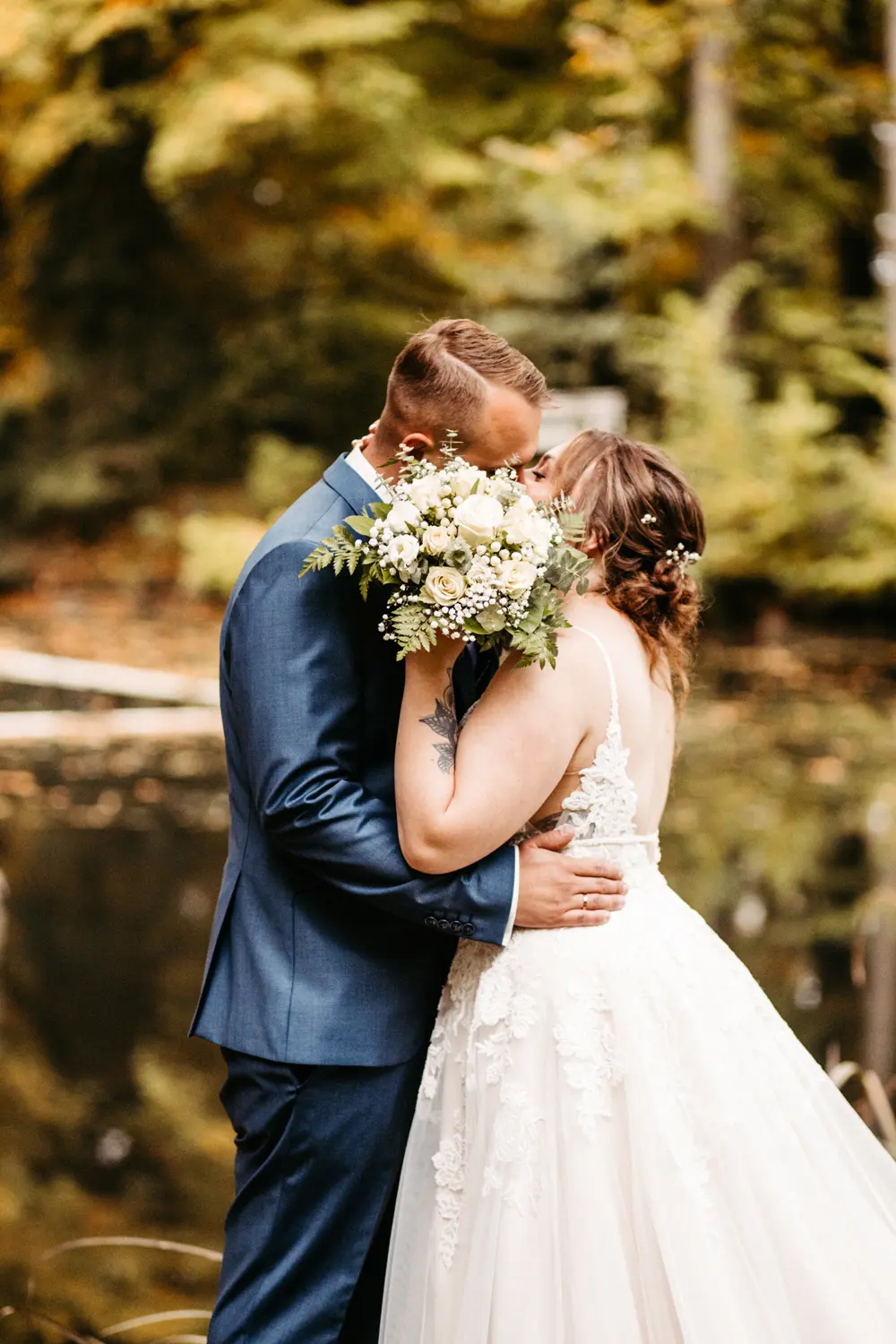 Eine Braut und ein Bräutigam küssen sich im Freien an einem Teich, wobei die Braut einen Strauß weißer Blumen vor ihr Gesicht hält. Sie sind umgeben von Grün und Herbstbäumen.