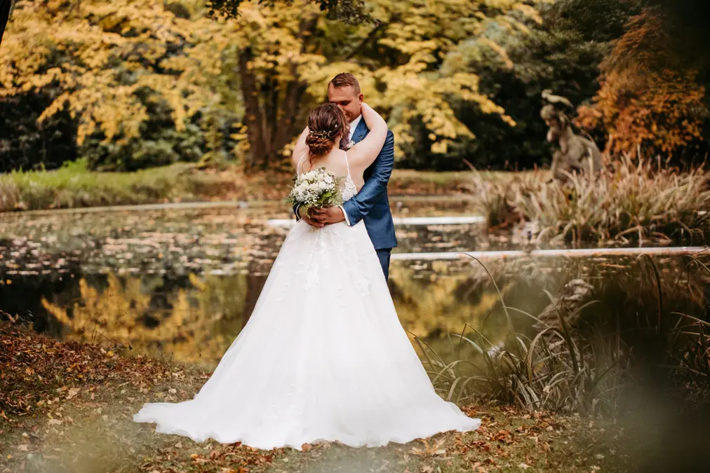Eine Braut und ein Bräutigam umarmen sich an einem Teich in einem Waldgebiet mit Herbstlaub. Die Braut trägt ein weißes Kleid und hält einen Blumenstrauß in der Hand, während der Bräutigam einen blauen Anzug trägt. Im Hintergrund ist eine Steinstatue zu sehen.