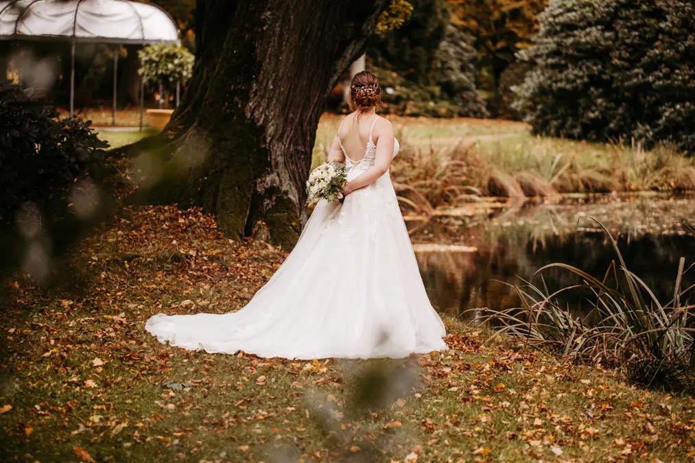 Eine Braut in einem weißen Hochzeitskleid steht im Freien an einem großen Baum und einem Teich und hält einen Blumenstrauß hinter ihrem Rücken. Der Boden ist mit Herbstlaub bedeckt und die Szene ist von Grün umgeben.