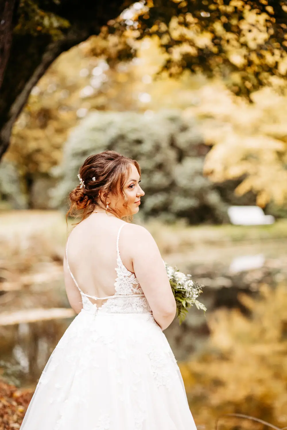 Eine Braut in einem weißen Spitzenhochzeitskleid steht im Freien in der Nähe eines Teiches und schaut über ihre Schulter. Sie hat rotes, mit Blumen hochgestecktes Haar und ist von Bäumen mit gelben und grünen Blättern umgeben.