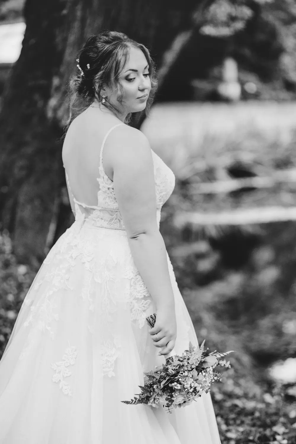 Eine Braut im Spitzenhochzeitskleid steht im Freien, hält einen Strauß in der Hand und blickt über ihre Schulter. Das Schwarz-Weiß-Foto zeigt sie in einer natürlichen, ruhigen Umgebung mit Bäumen und unscharfem Laub im Hintergrund.