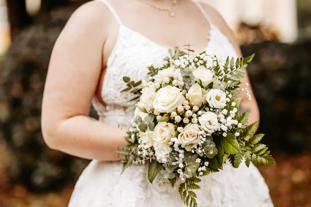 Eine Braut in einem weißen Spitzenkleid hält einen Strauß weißer Rosen, Grünzeug und Schleierkraut. Sie steht im Freien vor einem unscharfen natürlichen Hintergrund.
