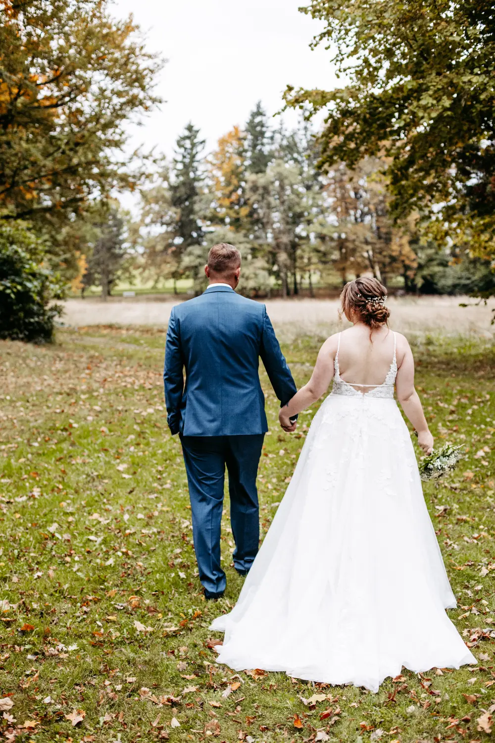 Eine Braut in einem weißen Kleid und ein Bräutigam in einem blauen Anzug gehen Hand in Hand über einen grasbewachsenen Weg im Freien, umgeben von Bäumen mit Herbstlaub, von hinten gesehen.
