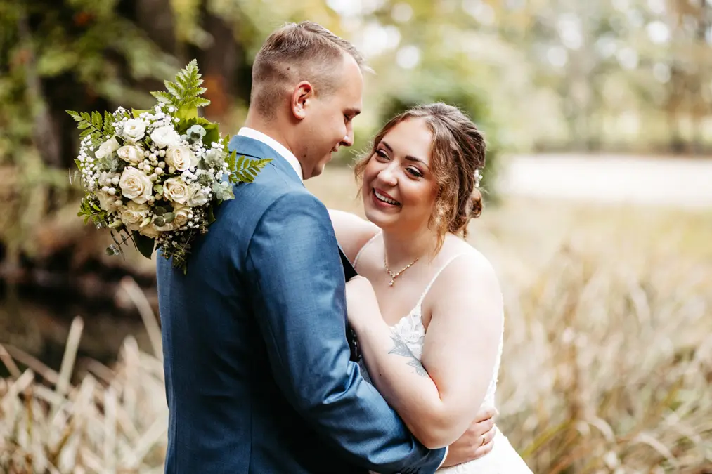 Eine Braut und ein Bräutigam umarmen sich im Freien und lächeln sich herzlich an. Der Bräutigam trägt einen blauen Anzug und hält einen Strauß aus weißen Blumen und Grünzeug. Die Braut trägt ein weißes Kleid. Der Hintergrund ist eine unscharfe natürliche Umgebung.