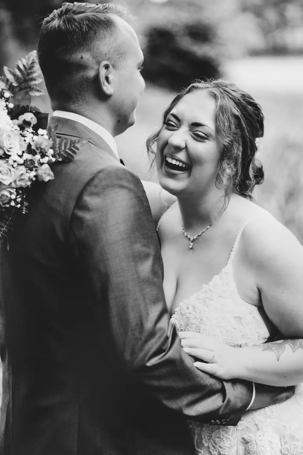Eine fröhliche Braut in einem Spitzenkleid lacht, während sie die Hände des Bräutigams hält, der ihr in einem Anzug gegenübersteht und einen Blumenstrauß in der Hand hält. Das Foto ist schwarz-weiß und fängt einen ehrlichen, glücklichen Hochzeitsmoment ein.