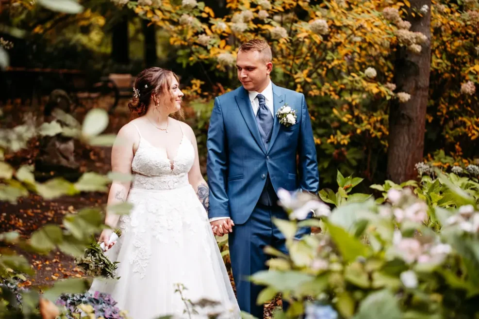 Eine Braut in einem weißen Kleid und ein Bräutigam in einem blauen Anzug halten sich an den Händen und lächeln sich an, während sie in einem üppigen, belaubten Garten mit Herbstlaub stehen.