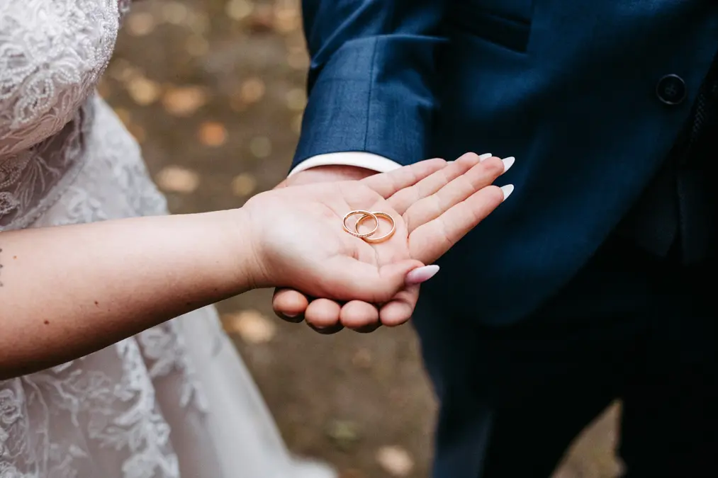 Eine Nahaufnahme einer Braut und eines Bräutigams, die sich an den Händen halten, wobei zwei goldene Eheringe auf der Handfläche der Braut ruhen. Die Braut trägt ein Spitzenkleid, der Bräutigam einen blauen Anzug.