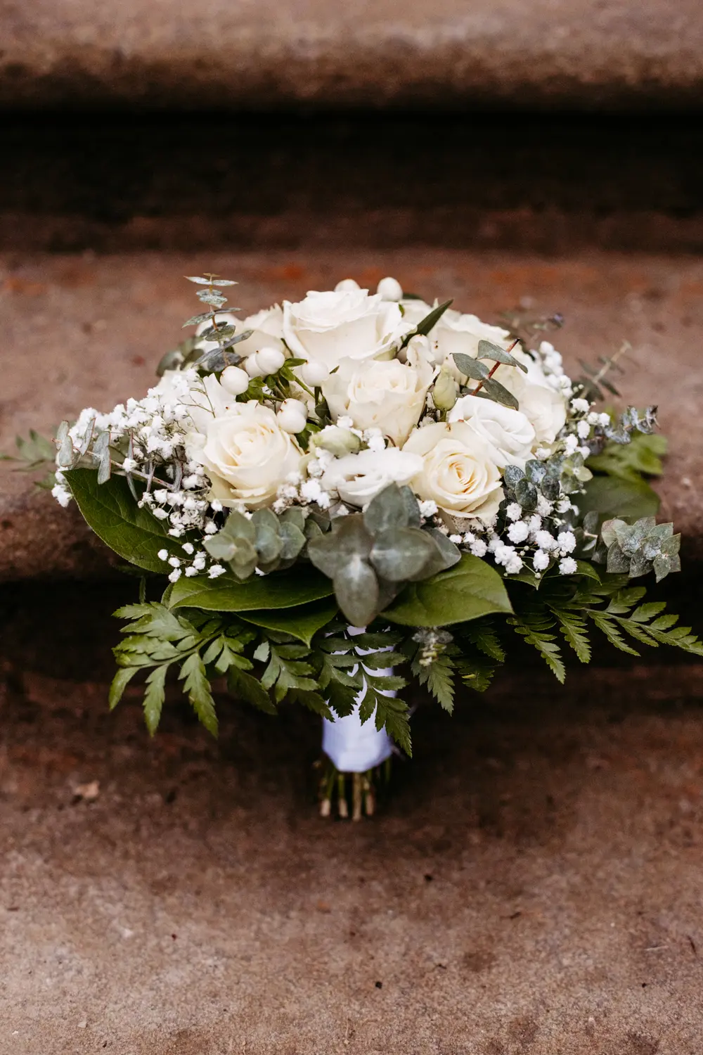 Ein BrautstrauĂź aus weiĂźen Rosen, Schleierkraut, Eukalyptus und grĂĽnen Farnen liegt auf einer Steinstufe.