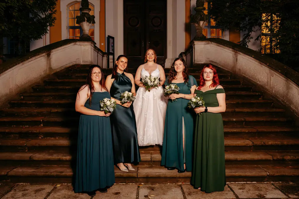 Eine Braut in einem weißen Kleid steht nachts auf einer Freitreppe, umgeben von vier Brautjungfern in verschiedenfarbigen grünen Kleidern, die alle kleine Sträuße halten und in die Kamera lächeln.