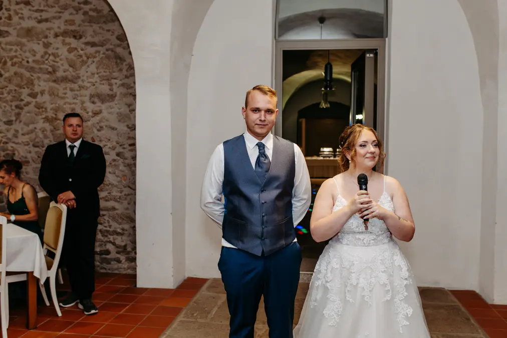 Eine Braut in einem weißen Kleid hält ein Mikrofon und steht neben einem Bräutigam in Weste und Krawatte. Sie stehen drinnen an einer Steinmauer, während die Gäste sitzen und ein Mann im Anzug im Hintergrund steht.