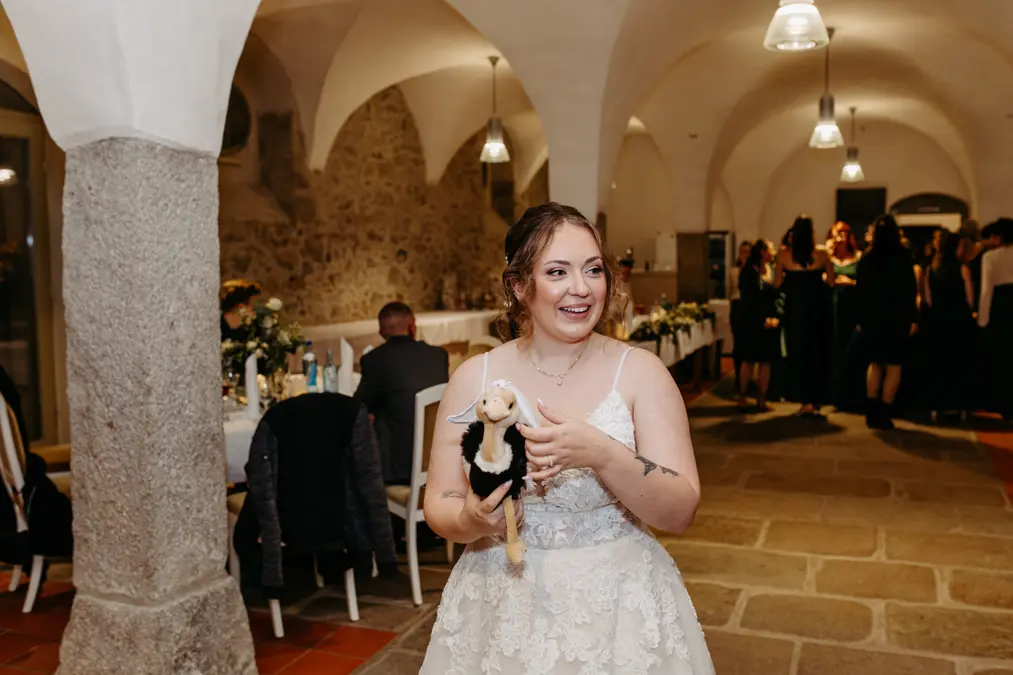 Eine lächelnde Braut in einem weißen Spitzenhochzeitskleid hält einen kleinen Plüschhund, während sie in einer geschmückten Empfangshalle mit sitzenden und stehenden Gästen im Hintergrund steht.