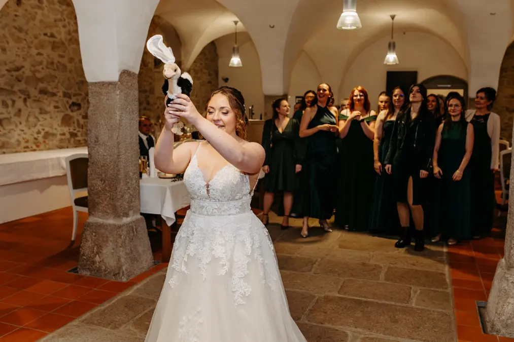 Eine Braut in einem weißen Hochzeitskleid bereitet sich darauf vor, den Brautstrauß einer Gruppe von Frauen zuzuwerfen, die hinter ihr in einem von Steinwänden umgebenen, gewölbten Raum mit warmer Beleuchtung stehen.