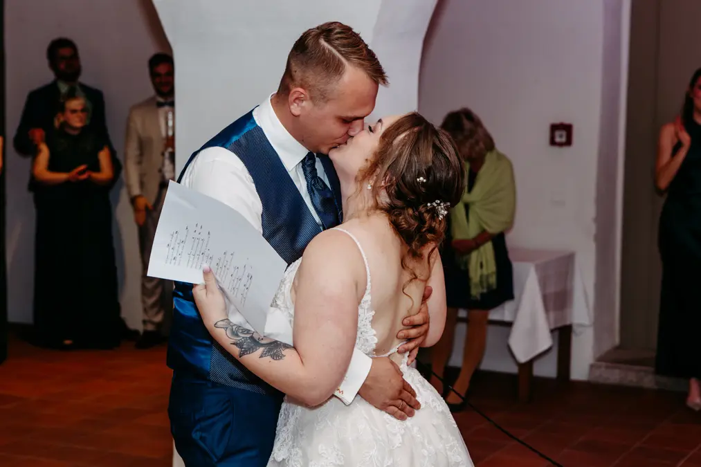 Braut und Bräutigam geben sich auf der Tanzfläche einen Kuss. Die Braut hält ein handgeschriebenes Schild in der Hand, und die Gäste stehen im Hintergrund und beobachten das Paar. Die Szene scheint bei einem Hochzeitsempfang im Haus zu spielen.