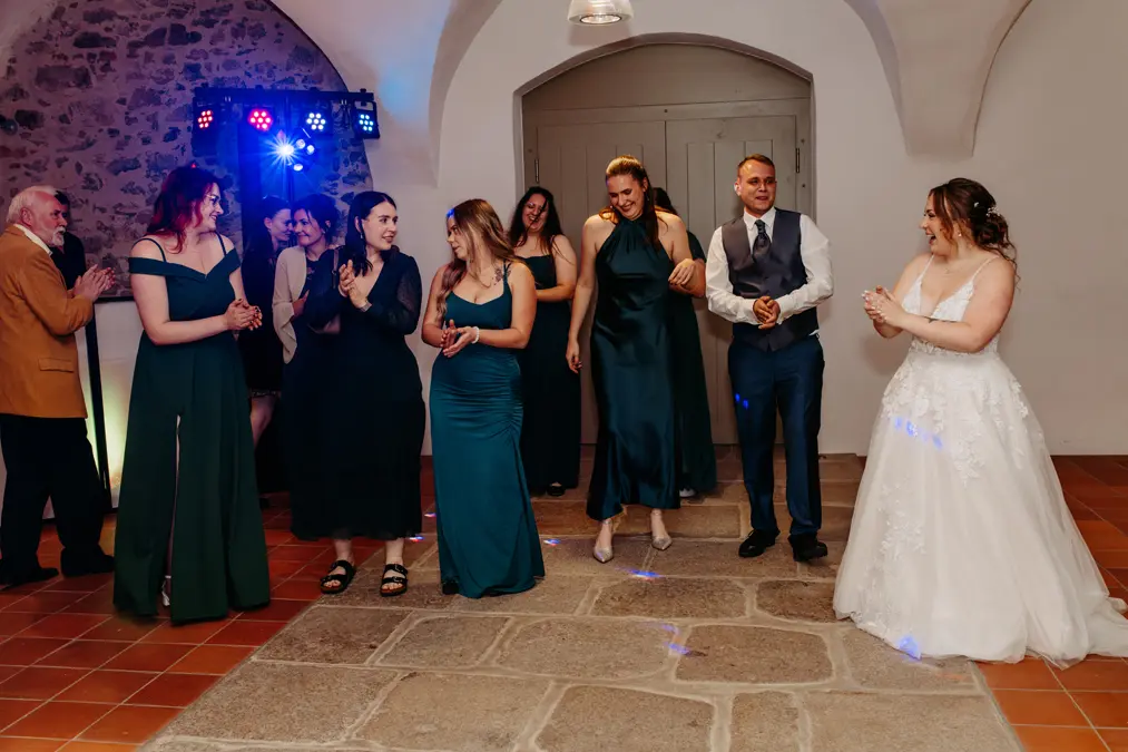 Eine Braut in einem weißen Kleid lächelt, während sie mit Brautjungfern in blaugrünen Kleidern und einem Trauzeugen im Anzug auf der Tanzfläche steht, umgeben von applaudierenden Gästen in einer warm beleuchteten Empfangshalle.