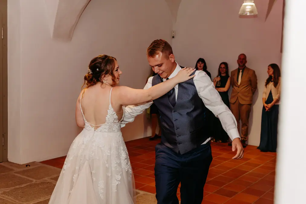 Eine Braut in einem weißen Kleid und ein Bräutigam mit Weste und Krawatte tanzen zusammen in einem warm beleuchteten Raum, während eine Gruppe von Gästen in formeller Kleidung aus dem Hintergrund zusieht.