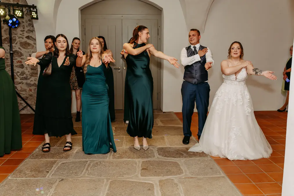 Fünf Personen, darunter vier Frauen in formellen Kleidern und ein Mann im Anzug, stehen in einem Innenraum auf einem Steinboden und führen einen Gruppentanz mit nach vorne und zur Seite gestreckten Armen auf, lächeln und amüsieren sich.