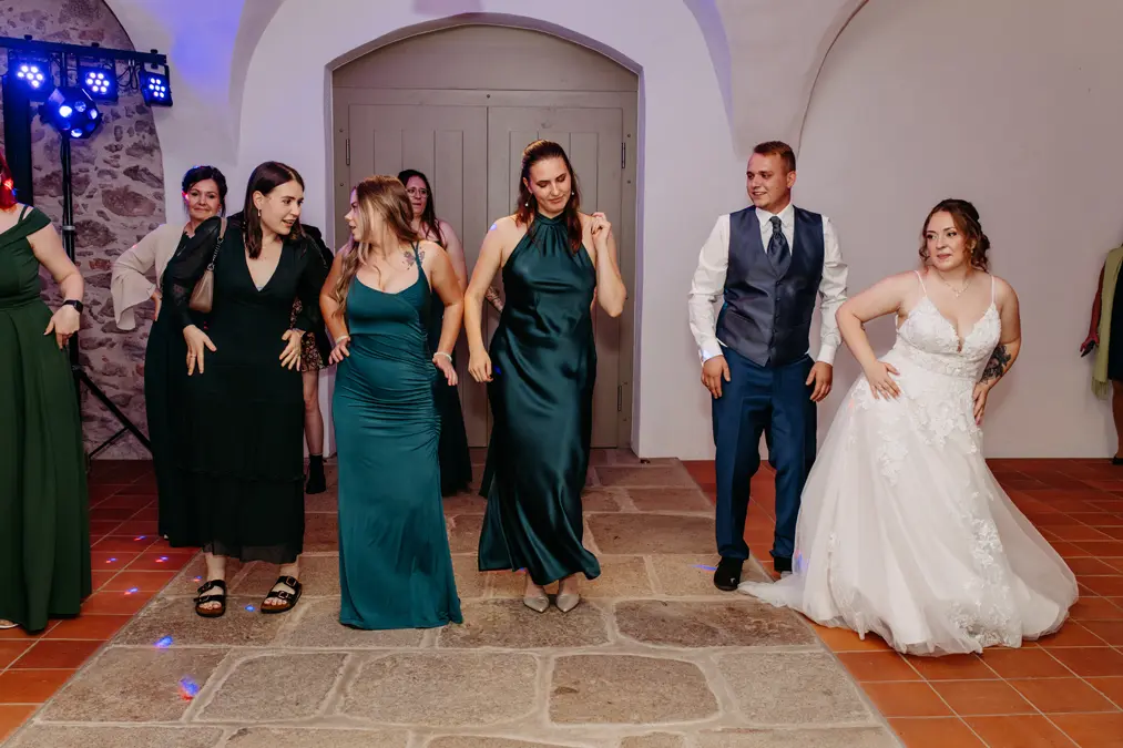 Eine Gruppe von Personen, darunter eine Braut in einem weißen Kleid und ein Mann in einer Weste, tanzt gemeinsam in einem Innenraum auf einem Steinboden. Andere Gäste in formeller Kleidung machen mit, lächeln und genießen den Moment.
