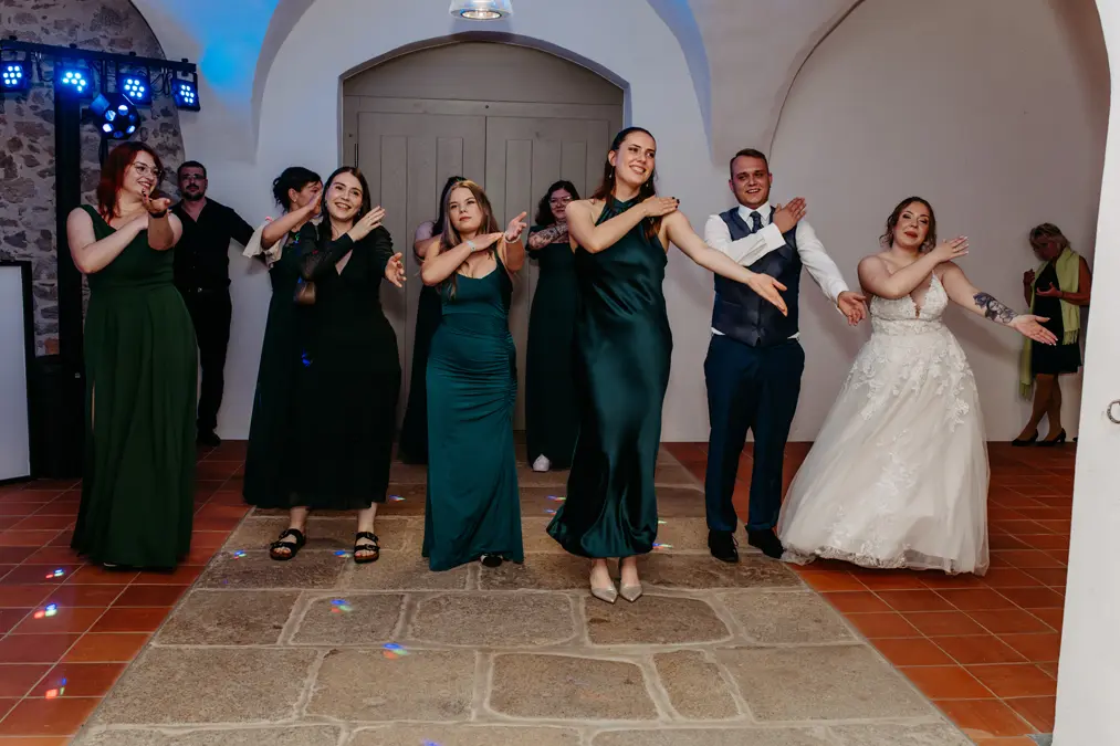 Eine Gruppe formell gekleideter Personen, darunter Brautjungfern in grünen Kleidern und eine Braut in einem weißen Kleid, tanzt in einem Innenraum auf einem Steinboden, lächelt und bewegt sich mit koordinierten Armgesten.