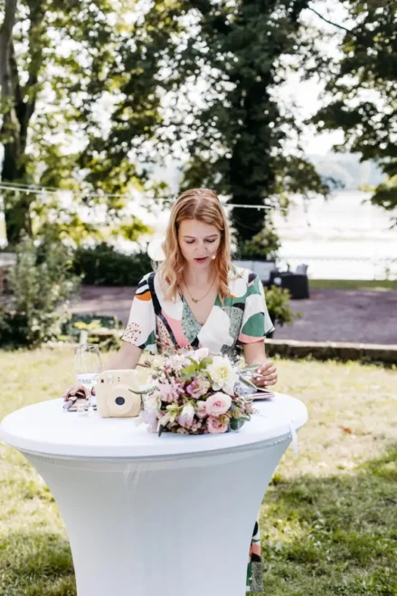 Die Traurednerin mit hellem Haar steht an einem kleinen runden Tisch im Freien. Sie trägt ein farbenfrohes Kleid und konzentriert sich auf die Aktivitäten vor ihr. Auf dem Tisch steht ein Blumenstrauß, im Hintergrund sind Bäume und Tische sanft verschwommen.