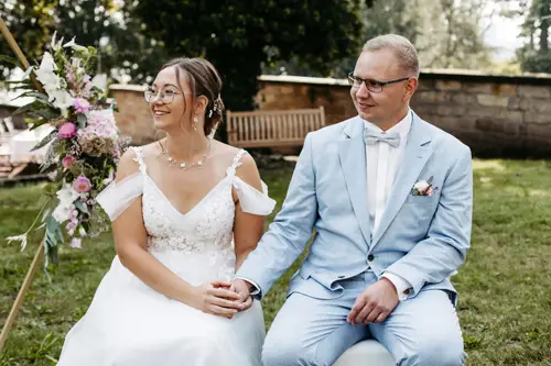 Braut und Bräutigam, gekleidet in ein weißes Spitzenbrautkleid und einen hellblauen Anzug, sitzen Händchen haltend auf Stühlen. Der Anzug des Bräutigams ist mit einem Einstecktuch und einer Ansteckblume geschmückt. Diese ruhige Zeremonie im Freien auf einer Wiese wurde von Melanie Kunert Fotografie, der renommierten Hochzeitsfotografin Dresden, wunderschön eingefangen.