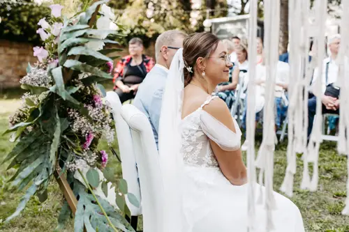 Eine Braut in einem weißen Kleid mit floraler Spitze sitzt im Freien neben einem Mann im hellen Anzug, fotografiert von einem Hochzeitsfotografen in Marcolinis Welt. Die Gäste sitzen auf Stühlen inmitten üppiger Vegetation und eleganter Blumenarrangements, während weiße Bänder anmutig neben ihnen hängen.