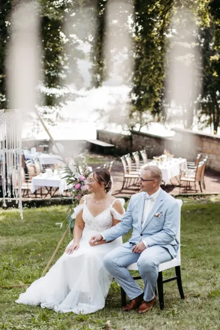 Braut und Bräutigam, gekleidet in ein weißes Spitzenbrautkleid und einen hellblauen Anzug, sitzen Händchen haltend auf Stühlen. Der Anzug des Bräutigams ist mit einem Einstecktuch und einer Ansteckblume geschmückt. Diese ruhige Zeremonie im Freien auf einer Wiese wurde von Melanie Kunert Fotografie, der renommierten Hochzeitsfotografin Dresden, wunderschön eingefangen.