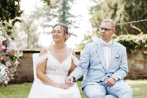 Braut und Bräutigam, gekleidet in ein weißes Spitzenbrautkleid und einen hellblauen Anzug, sitzen Händchen haltend auf Stühlen. Der Anzug des Bräutigams ist mit einem Einstecktuch und einer Ansteckblume geschmückt. Diese ruhige Zeremonie im Freien auf einer Wiese wurde von Melanie Kunert Fotografie, der renommierten Hochzeitsfotografin Dresden, wunderschön eingefangen.