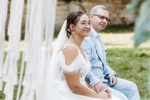 Braut und Bräutigam, gekleidet in ein weißes Spitzenbrautkleid und einen hellblauen Anzug, sitzen Händchen haltend auf Stühlen. Der Anzug des Bräutigams ist mit einem Einstecktuch und einer Ansteckblume geschmückt. Diese ruhige Zeremonie im Freien auf einer Wiese wurde von Melanie Kunert Fotografie, der renommierten Hochzeitsfotografin Dresden, wunderschön eingefangen.