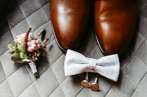 Braune Lederschuhe, eine hellgraue Fliege und passende Manschettenknöpfe sind elegant auf einer gesteppten grauen Oberfläche arrangiert. Eine kleine Ansteckblume mit rosa und weißen Blumen verleiht der Komposition von Hochzeitsfotografin Dresden – Melanie Kunert Fotografie einen besonderen Charme.