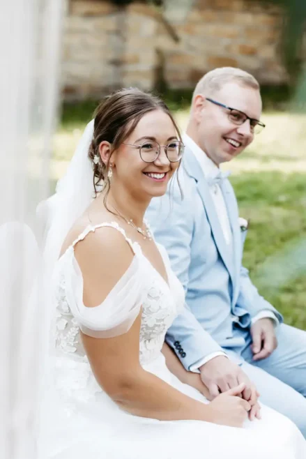 Braut und Bräutigam, gekleidet in ein weißes Spitzenbrautkleid und einen hellblauen Anzug, sitzen Händchen haltend auf Stühlen. Der Anzug des Bräutigams ist mit einem Einstecktuch und einer Ansteckblume geschmückt. Diese ruhige Zeremonie im Freien auf einer Wiese wurde von Melanie Kunert Fotografie, der renommierten Hochzeitsfotografin Dresden, wunderschön eingefangen.