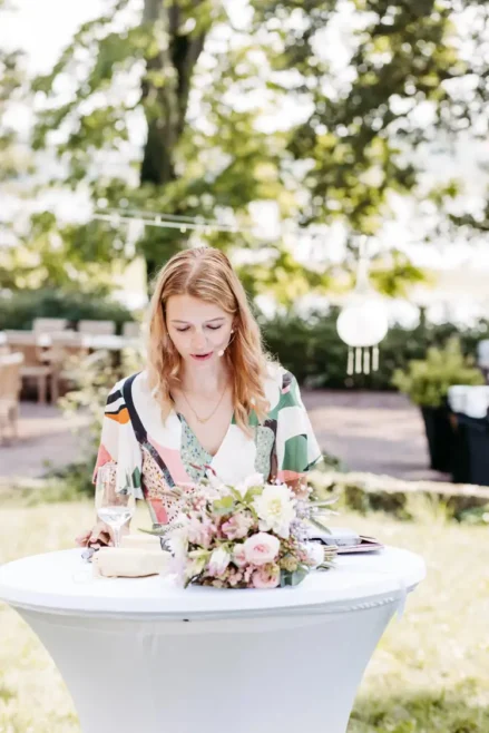 Die Traurednerin mit hellem Haar steht an einem kleinen runden Tisch im Freien. Sie trägt ein farbenfrohes Kleid und konzentriert sich auf die Aktivitäten vor ihr. Auf dem Tisch steht ein Blumenstrauß, im Hintergrund sind Bäume und Tische sanft verschwommen.