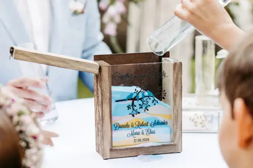 Eine Holzkiste mit der Gravur „Together We Make a Family“ auf dem Deckel enthält eine dekorative Glasplatte mit einem Baummotiv und Daten. In der nahegelegenen Marcolinis Welt nehmen Gäste an einer Sandzeremonie für eine Hochzeit teil, die vom erfahrenen Blick des Hochzeitsfotografen festgehalten wurde.