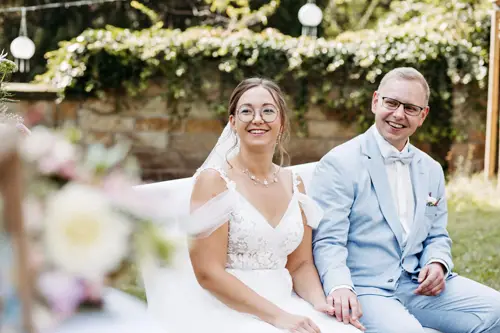 Eine Braut im weißen Kleid und ein Bräutigam im hellblauen Anzug sitzen lächelnd und Händchen haltend im Freien. Umgeben von Grün und mit Lichterketten beleuchteten Steinmauern wirkt die Szene hell und fröhlich – eine perfekte Aufnahme von Melanie Kunert Fotografie, Hochzeitsfotografin Dresden.
