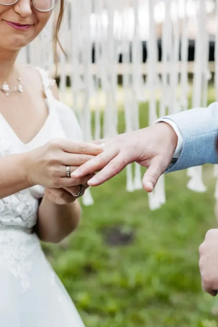 Bei einer Hochzeit, die von Hochzeitsfotograf Dresden fotografiert wurde, steckt die Braut dem Bräutigam elegant einen Ring an den Finger. Sie glänzt in einem weißen Kleid mit Spitzendetails, während er einen hellblauen Anzug trägt. Dieser bezaubernde Moment entfaltet sich vor einer mit weißen Dekorationen geschmückten Außenkulisse.