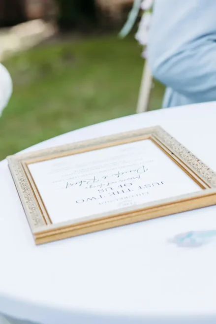 Ein gerahmtes Schild liegt flach auf einem runden weißen Tisch im Freien, möglicherweise von einem Hochzeitsfotografen in Marcolinis Welt aufgestellt. Das Schild mit teilweise unleserlichem Text liegt auf dem Gras, während eine Person in einem hellblauen Outfit daneben steht und dieser malerischen Szene Charme verleiht.