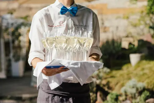 Eine Person in weißem Hemd und blauer Fliege balanciert gekonnt ein Tablett mit mehreren Gläsern Weißwein oder Champagner, elegant eingefangen von einem Hochzeitsfotografen in Marcolinis Welt vor einer unscharfen Außenkulisse.
